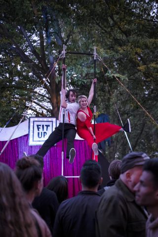 A photograph of two artists performing aerial tricks. They hold themselves high above the audience. One performer wears a red dress, the other performer wears a white shirt and black trousers.