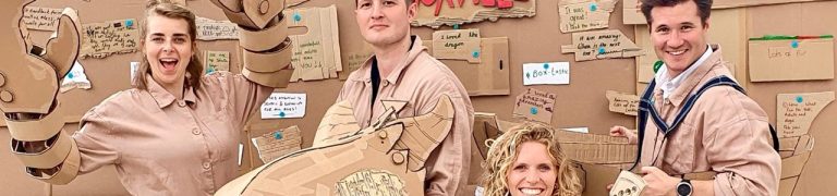 Performers stand wearing brown combat suits, the make a performance from cardboard.