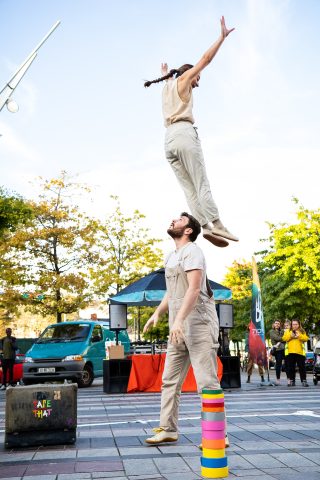 Two performers wear beige clothing and perform infront of an audience. One is leaping in the air whilst the other looks up towards them. There are colourful rolls of duct tape stacked as part of the performance.