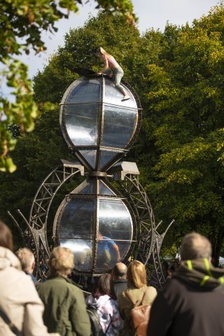 A performer pulls against a large metal sandtimer. Green trees are out of focus in the background.