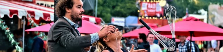 Two street artists perform in the middle of a festival. One performer stands behind the other pouring water into their mouth while they juggle with kitchen whisks.