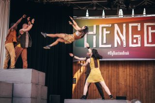 A performance of HENGE. One performer jumps from a stack of grey stones into the arms of two other performers.