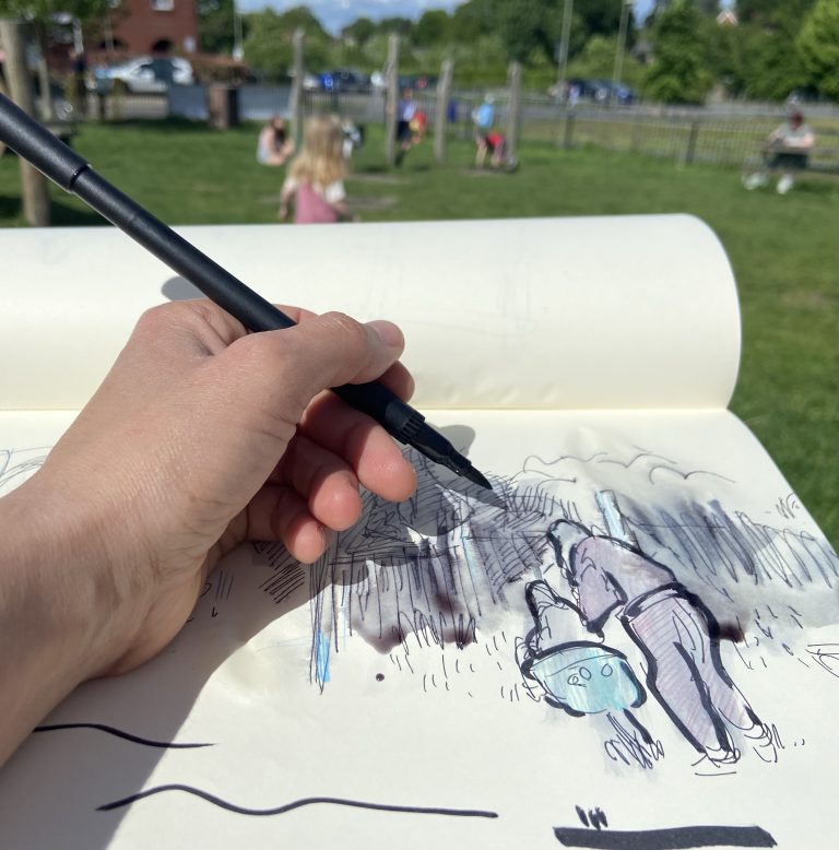 A close up photograph of a persons hand drawing in a sketchbook. The person uses a black pen on the paper. In the backdrop is a blurred playground with children playing.