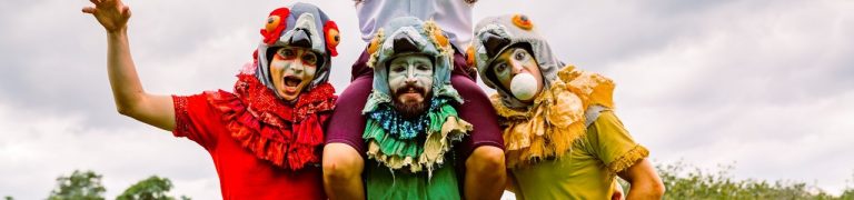 Three performers dress up as birds, they stand in a huddle. Each performer has a headpiece, one in red, one in yellow and one in green.