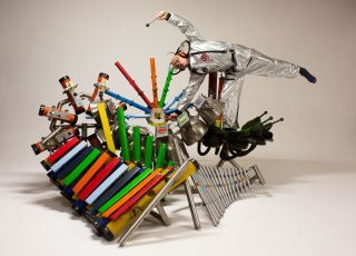 A performer wears a silver suit and performs a cartwheel on top of musical instruments made from a variety of objects.