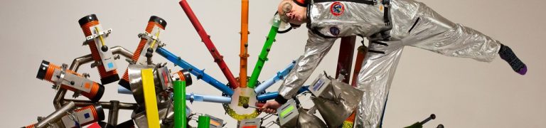 A performer wears a silver suit and performs a cartwheel on top of musical instruments made from a variety of objects.