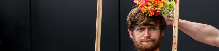 A photograph of the performance SPLATT! A character looks through a wooden frame with a glum face. An arm of another performer places a bunch of flowers on their head. The background is black.