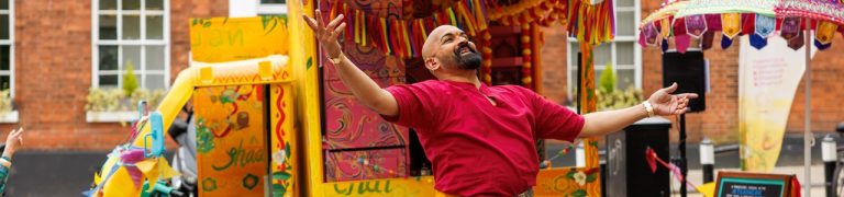 A performer stands with their arms open wide looking to the sky. They wear a vivid red t-shirt and stand infront of a bright yellow TukTuk.