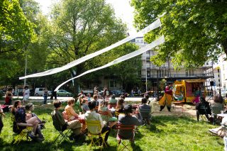 A performance outside in a park. An audience watch on as two performers dance infront of a yellow TukTuk.