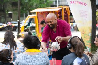 A photograph of an outdoor performance. Shows a person in a red top pouring a cup of tea for a member of the audience. He smiles. In the background is a bright yellow tuktuk.