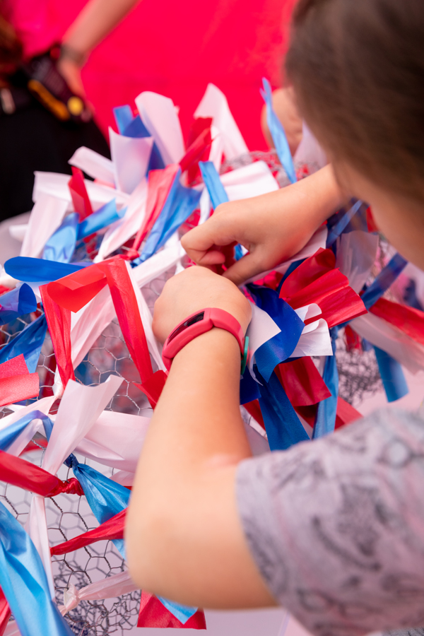 A close up image shows a pair of childs hands trying red, blue and white ribbons to a wire structure.