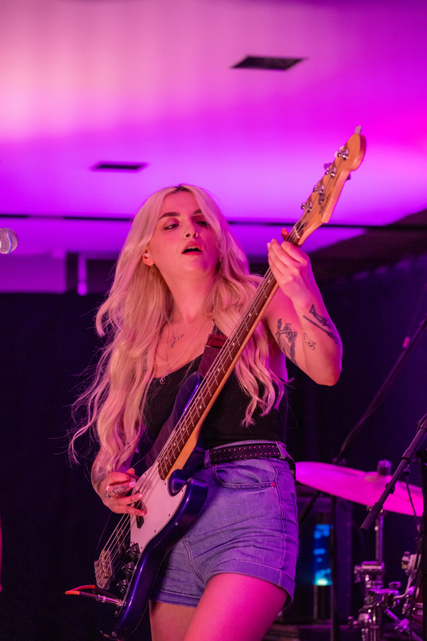 A guitarist playing a black electric guitar. They have long blonde hair and wear a jean skirt. The lighting is purple.