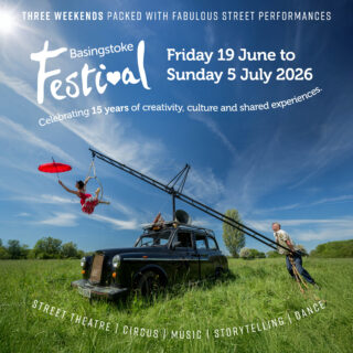 Two performers use a black taxi as part of a performance. The taxi is parked on bright green grass on a sunny day. One performer wears red and hangs from the top of a black ladder which is fixed to the roof of the taxi. The second performer stands on the grass holding the base of the ladder. Large white logo Basingstoke Festival white text, Friday 19 June to Sunday 5 July 2026. Three weekends packed with fabulous street performances. Celebrating 15 years of creativity, culture and shared experiences.