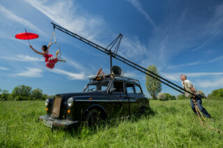 Two performers use a black taxi as part of a performance. The taxi is parked on bright green grass on a sunny day. One performer wears red and hangs from the top of a black ladder which is fixed to the roof of the taxi. The second performer stands on the grass holding the base of the ladder. Large white logo Basingstoke Festival white text, Friday 19 June to Sunday 5 July 2026. Three weekends packed with fabulous street performances. Celebrating 15 years of creativity, culture and shared experiences.