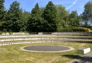 A tarmac circle with grass around and then steps that are seating. You can see trees and a blue sky in the background.