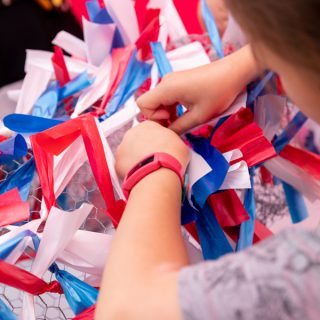A close up image shows a pair of childs hands trying red, blue and white ribbons to a wire structure.