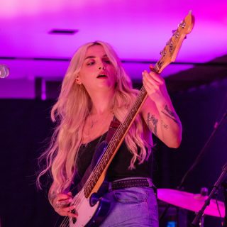 A guitarist playing a black electric guitar. They have long blonde hair and wear a jean skirt. The lighting is purple.